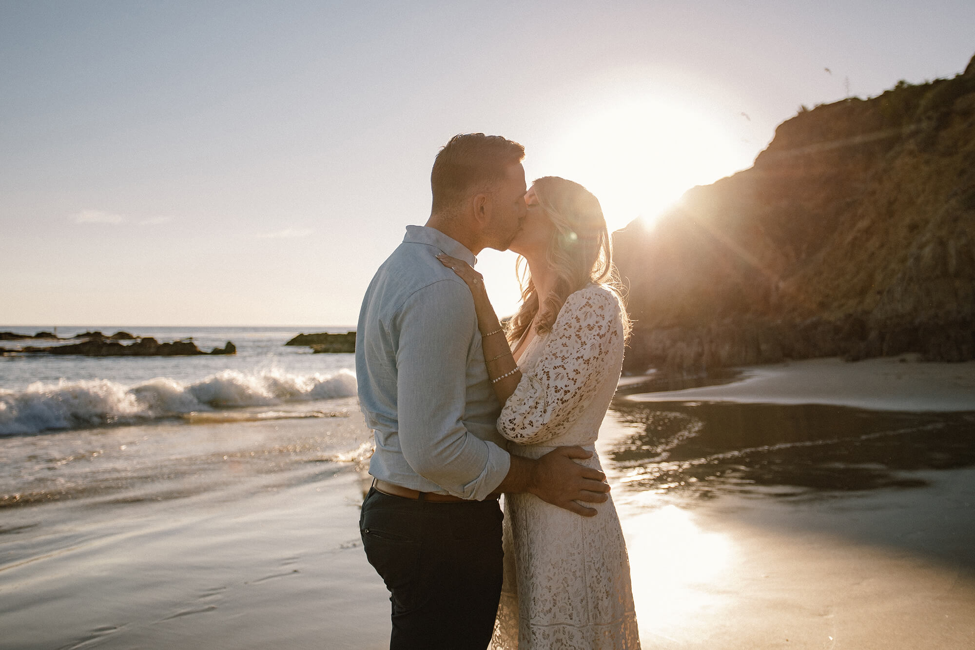 JJ and Kait kissing on the beach with the sunset behind them.
